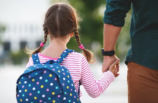 Ein Mädchen geht mit einem Erwachsenen Hand in Hand und trägt einen Rucksack